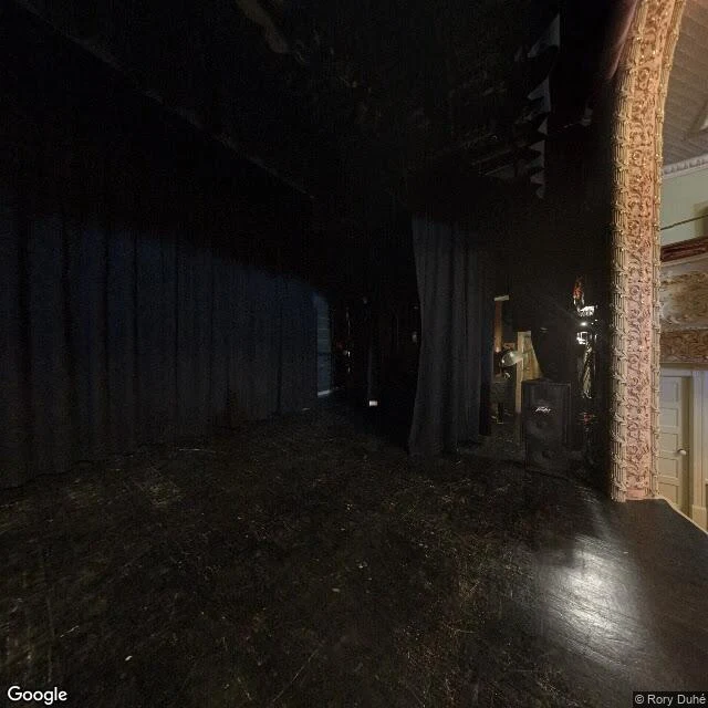 Interior of the Haskell Opera House stage: worn dark stage floor, black curtains, an ornate carved column, and stage lighting.