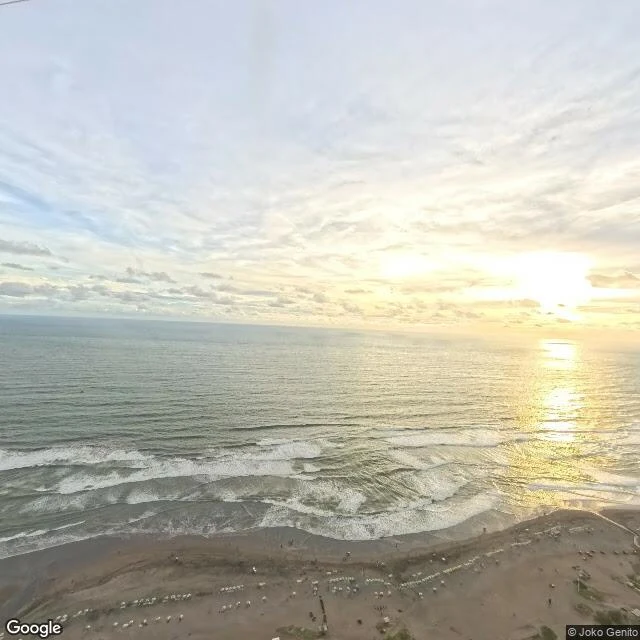 The south coast from the air: a low sun lays a shining path across the Indian Ocean; umbrellas stand on pale sand like white seeds. Frame by Joko Genito, December 2025.
