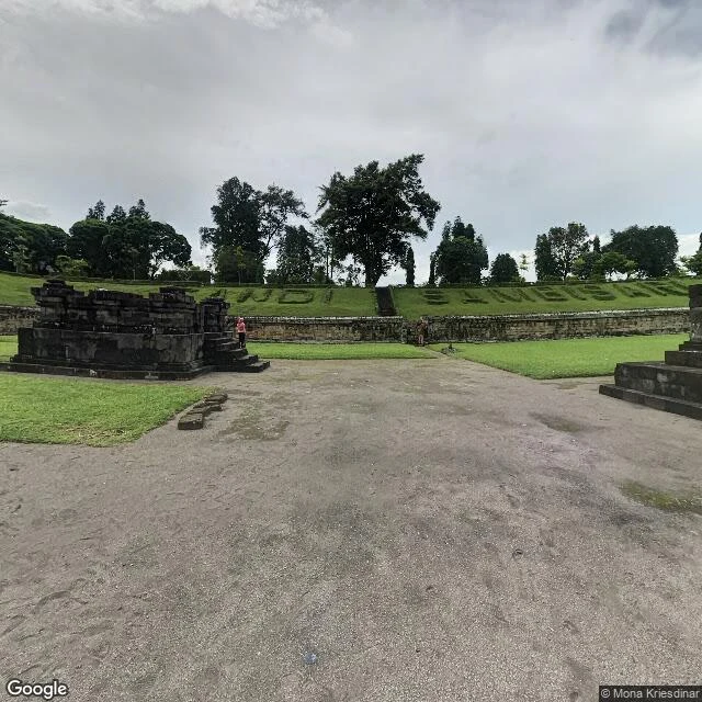 A visitor in pink stands on the gravel in the court of Sambisari, with the temple bank above her. Street view, July 2016.